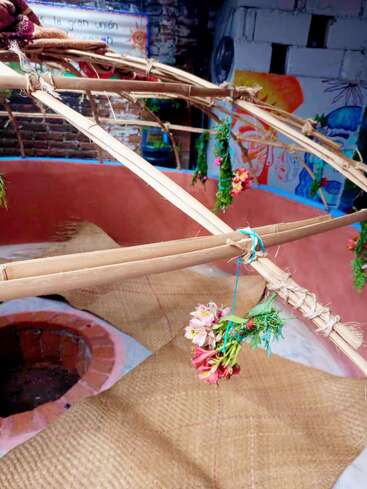 This image shows a traditional sweat lodge structure with bamboo poles, woven mats, flowers, and greenery tied inside. Colorful murals and brick wall are visible behind.