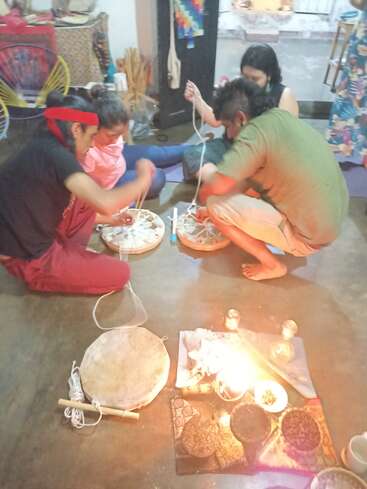 Four people sit on the floor making traditional drums, surrounded by candles and offering bowls, creating a warm, communal, and spiritual craft atmosphere indoors.