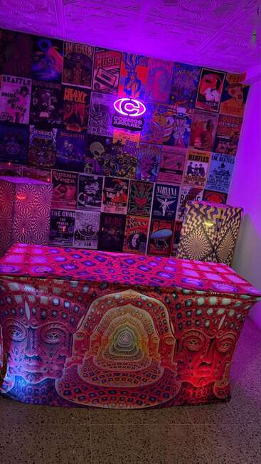 Una habitación con temática psicodélica cuenta con vívidos pósters en las paredes, un letrero de neón brillante y una mesa cubierta con un tapiz vibrante con ilustraciones surrealistas y alucinantes de caras iluminadas por luces moradas.