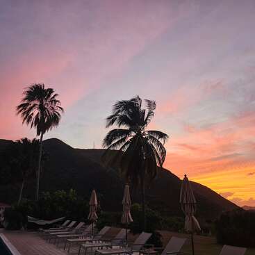 Palmen vor einem leuchtenden Sonnenuntergang, Berge im Hintergrund, Liegestühle und Sonnenschirme am Pool schaffen eine heitere und tropische Urlaubsatmosphäre.