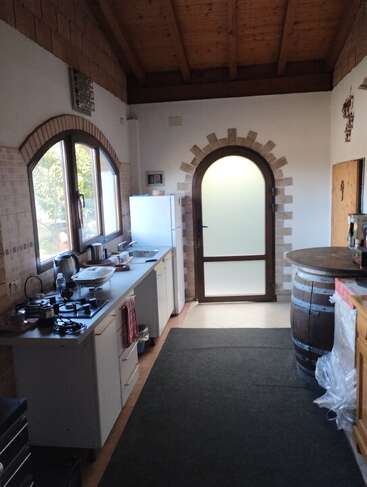 Cette cuisine rustique est dotée de fenêtres et de portes cintrées, de poutres apparentes, d'une table en tonneau de vin, d'armoires blanches, d'un réfrigérateur et d'ustensiles de cuisine indispensables sur le plan de travail.