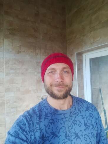 Un homme barbu portant un bonnet rouge en tricot et un pull bleu à motifs se tient debout à l'intérieur, souriant. L'arrière-plan montre des murs carrelés de couleur beige et une porte vitrée.