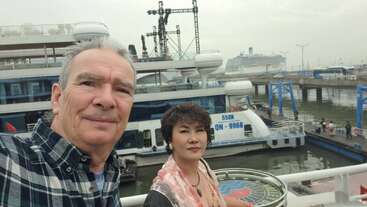 Um homem e uma mulher tiram uma selfie em um cais, com barcos e um navio de cruzeiro ao fundo, sob um céu nublado. Cena movimentada do porto.