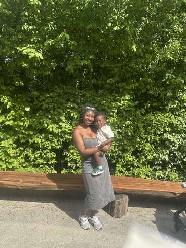 A smiling woman in a grey dress holds a young child. They stand in front of green bushes, with a wooden bench and sunlight illuminating the scene.