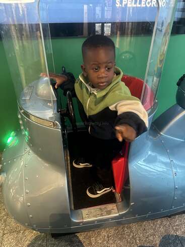 A young boy stands inside a coin-operated ride shaped like a vehicle. He looks to his right, pointing, wearing a green and black jacket with sneakers.