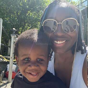 A joyful woman wearing sunglasses and a smiling young child pose together outdoors on a sunny day, surrounded by greenery and bright, natural light.