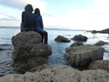 Two people sit on a large rock in the water, surrounded by smaller rocks, with a vast body of water and a cloudy sky in the background.