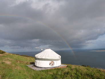 The image depicts a white yurt situated on a wooden platform, overlooking a vast body of water, with a rainbow stretching across the sky above.