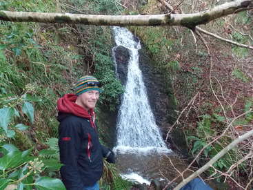 The image depicts a man standing in front of a small waterfall in a wooded area, surrounded by lush greenery and branches.