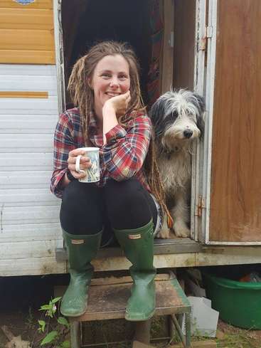 A woman with dreadlocks sits on a step outside a camper, holding a mug, with a dog standing beside her, both looking at the camera.