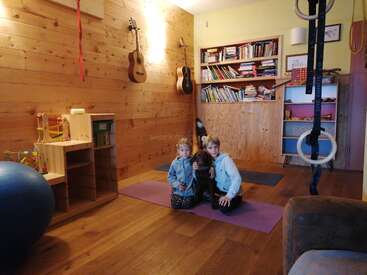 Two children and a dog sit on yoga mats in a cozy, wooden room with bookshelves, hanging guitars, gym rings, toys, and an exercise ball visible.