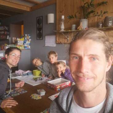 A family of five sits around a wooden table playing a board game. They smile warmly, surrounded by cozy decor, games, and a relaxed, homey atmosphere.