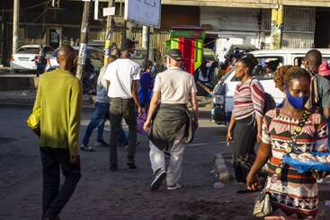 People walk through a busy urban street, some wearing face masks. Vehicles, shops, and a brightly colored kiosk are visible. The atmosphere is lively and bustling.