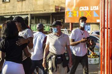 Several people walk outdoors in a busy urban setting. Most wear face masks and casual clothes. There is a colorful "Casa Lisa" sign in the background.