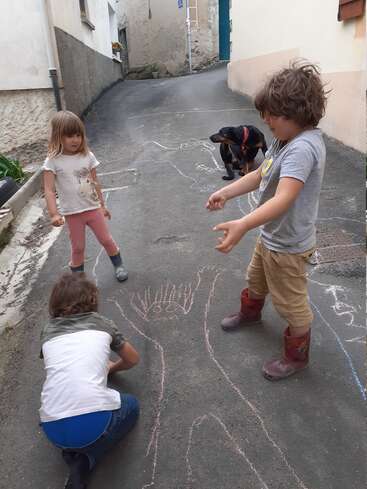 The image shows three children and a dog playing with chalk on a street, drawing on the pavement in a residential area.