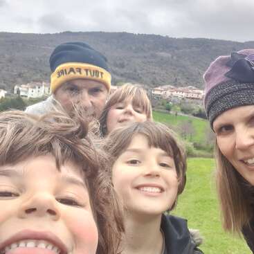 A imagem mostra uma família de cinco pessoas posando para uma selfie em um campo gramado com casas e montanhas ao fundo em um dia nublado.
