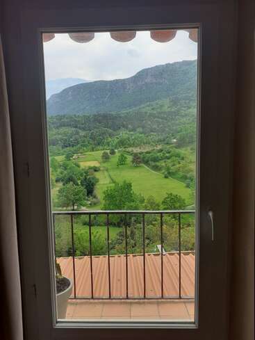 The image depicts a serene view of a mountainous landscape through a window, featuring a balcony with a railing and a potted plant, surrounded by lush greenery.