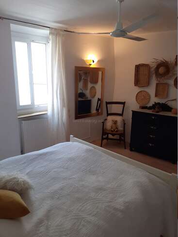 The image depicts a serene bedroom with a white bed, a dark dresser, and a mirror, featuring a white ceiling fan and a window with a white curtain.