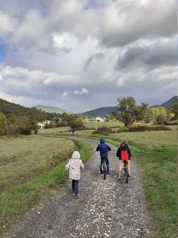 Three children ride bikes on a gravel path through a grassy field, with houses and trees in the background, under a cloudy sky.