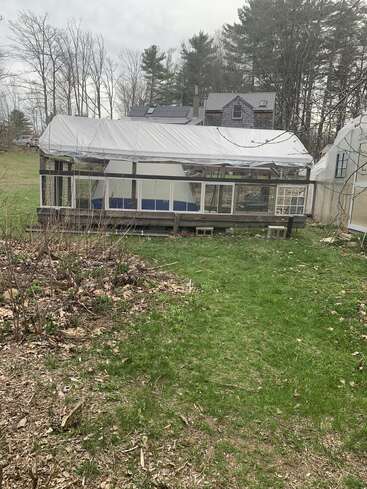 A covered greenhouse structure sits in a grassy backyard. Behind it, there’s a rustic house with solar panels. Surrounding trees are mostly leafless, hinting early spring.