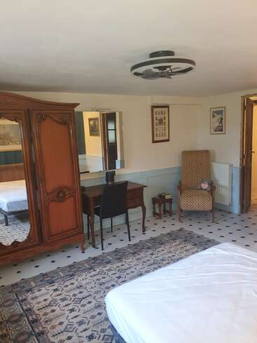Esse quarto tem um guarda-roupa clássico de madeira, uma mesa com cadeira, poltrona estampada, mesinha lateral com livros, espelhos, quadros, tapete grande e piso de azulejo.