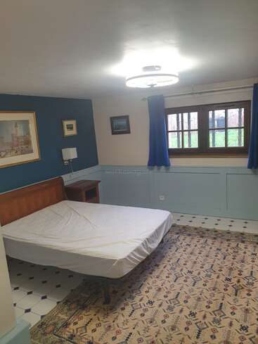 Esse é um quarto simples com uma cama de solteiro, tapete estampado, cortinas azuis, quadros nas paredes, piso de azulejo e janela de madeira. Ambiente aconchegante.