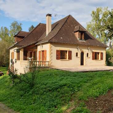 A charming country house with a steep, dark roof, arched doorway, wooden shutters, beige walls, surrounded by lush greenery, trees, and a cloudy sky.