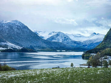 A serene lake reflects the surrounding snow-capped mountains. Green grass in the foreground is dusted with snow, while scattered homes nestle peacefully along the shoreline.
