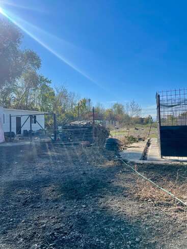 Una zona exterior iluminada por el sol con un suelo de tierra, neumáticos esparcidos, vallas metálicas, palés apilados, árboles, un edificio blanco y un cielo azul con rayos de sol.