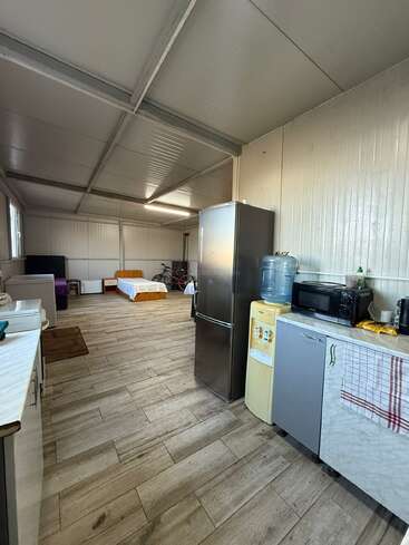 Esta imagen muestra una habitación sencilla y espaciosa con zona de cocina, frigorífico, dispensador de agua, microondas, cama, sofá, suelo de madera y bicicletas al fondo.