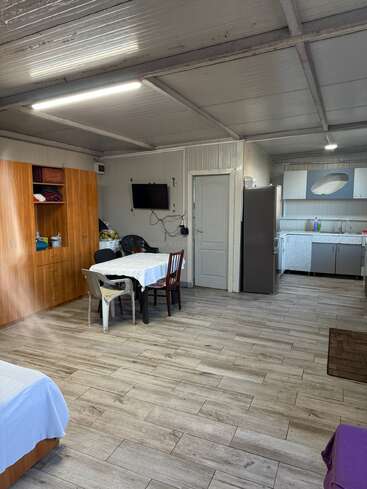 The image shows a simple, tidy room with a wardrobe, dining table with mixed chairs, a TV, fridge, kitchen area, tiled floor, and bright lighting.