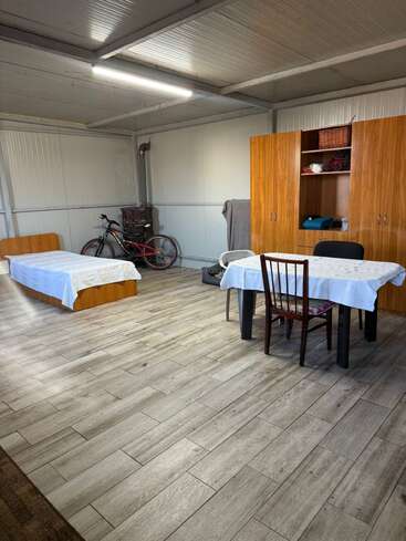 This room contains a single bed, table with chairs, wardrobe, bicycle, and wood stove. The floor is tiled, and the setting looks simple and functional.