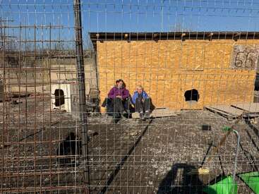 Two people sit beside a wooden shelter inside a fenced enclosure, possibly an animal shelter or rescue. The area is sunny and appears to be outdoors, somewhat barren.
