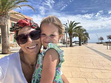 Una mujer y una niña sonríen y se abrazan mientras se hacen un selfie al aire libre en un día soleado. Las palmeras, el cielo azul y los edificios crean un alegre ambiente vacacional.