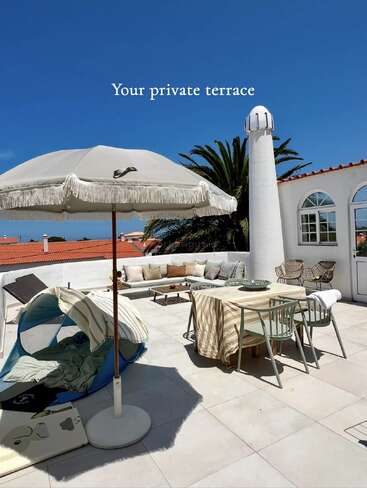 Une terrasse privée ensoleillée avec un grand parasol, un coin salon extérieur, une table à manger, des chaises longues, un palmier et un ciel bleu éclatant. Une atmosphère accueillante et relaxante.