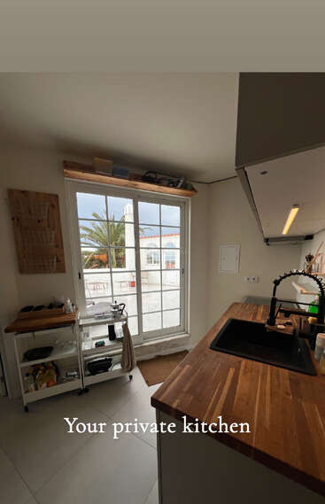 Une cuisine moderne et confortable avec des plans de travail en bois, un évier noir et une lumière abondante provenant des grandes portes vitrées donnant sur une terrasse avec des palmiers à l'extérieur.