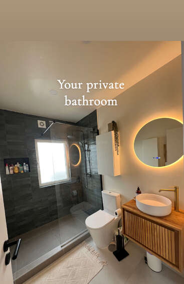 Salle de bain moderne avec douche carrelée sombre, porte vitrée, fenêtre, miroir circulaire rétroéclairé, toilettes blanches, meuble-lavabo en bois, lavabo vasque, articles de toilette soigneusement rangés, décoration minimaliste, éclairage tamisé.
