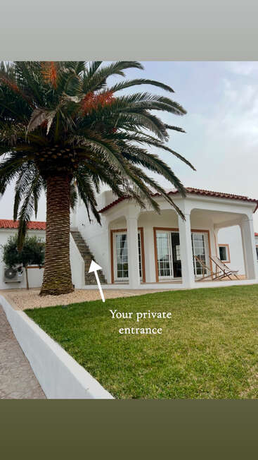 Une belle villa blanche avec un toit en terre cuite, un palmier devant, une pelouse verte, des escaliers en pierre et une entrée privée signalée menant à l'étage supérieur.