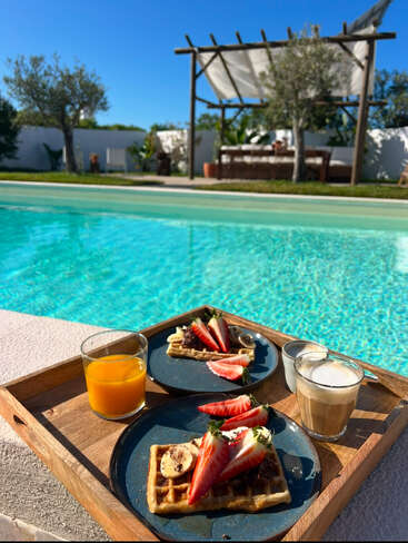 Un plateau en bois contient des gaufres garnies de fraises et de bananes, du jus d'orange et du café, au bord de la piscine, avec un ciel bleu éclatant et une pergola en arrière-plan.