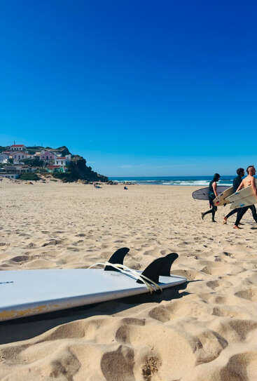 Une plage ensoleillée sous un ciel bleu limpide, une planche de surf sur le sable, des surfeurs marchant vers l'océan et des maisons colorées sur une falaise. Une atmosphère relaxante.