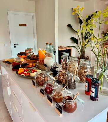 Un buffet petit-déjeuner copieux comprend des fruits frais, des œufs, du muesli, du jus, du lait, des confitures, des noix et des pâtes à tartiner dans des bocaux en verre, disposés sur un comptoir de cuisine moderne.