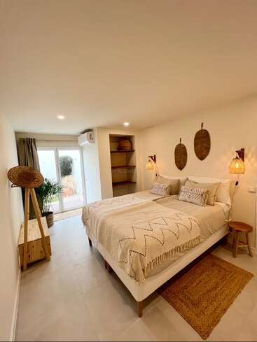 Cette chambre dispose d'un lit double confortable avec une literie beige, d'une décoration en osier, de meubles en bois, d'étagères murales, d'un éclairage naturel, de plantes d'intérieur et de portes coulissantes donnant sur l'extérieur.