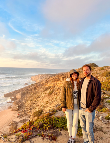 Un couple souriant se tient sur une falaise surplombant l'océan au coucher du soleil. Le ciel est partiellement nuageux et les vagues viennent se briser contre la côte accidentée et pittoresque en contrebas.