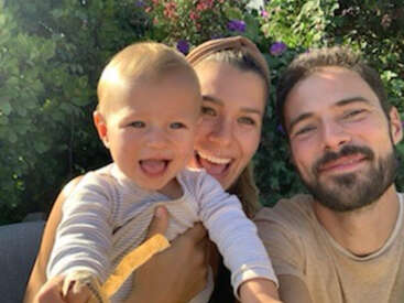 Eine glückliche dreiköpfige Familie, bestehend aus einem lächelnden Baby, einer jungen Frau und einem bärtigen Mann, die draußen ein fröhliches Selfie machen, während im Hintergrund Blumen blühen.