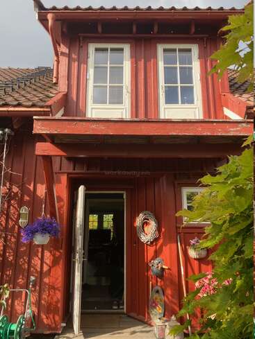 This image shows a charming red wooden house with white-trimmed windows and an open door. Flower pots and garden tools decorate the cozy, sunlit entrance.