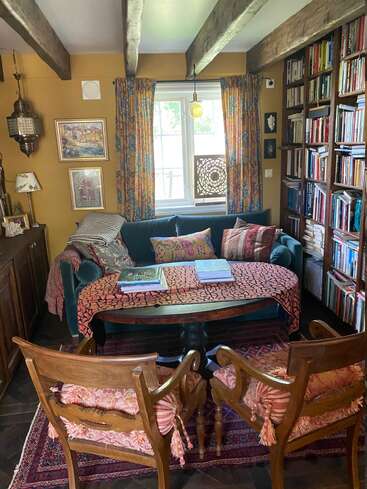 This cozy reading nook features a teal sofa, patterned pillows, a wooden table, two cushioned chairs, bookshelves, vibrant curtains, and warm, inviting decor accents.
