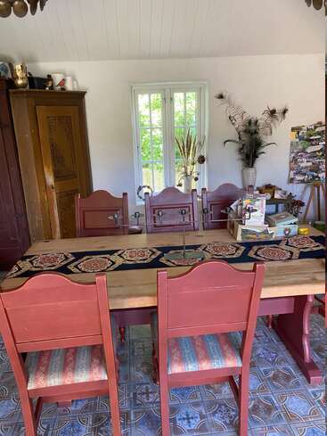 A cozy dining room features red wooden chairs, a patterned table runner, a large wooden wardrobe, window with greenery outside, dried plant arrangements, and colorful tiled floor.