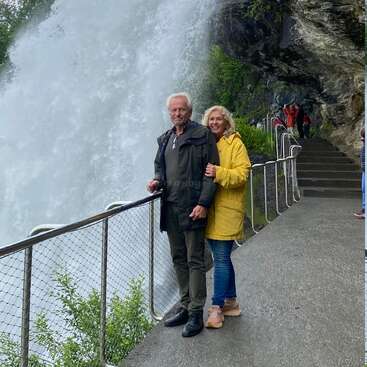 An older couple stands happily near a powerful waterfall, smiling and hugging. They wear raincoats, standing on a wet path with stairs and railings nearby.