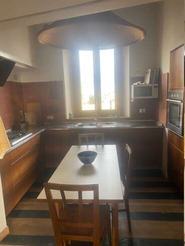This cozy kitchen features wooden cabinets, a central table with two chairs, a black bowl, large window, woven light fixture, microwave, stove, oven, and striped flooring.