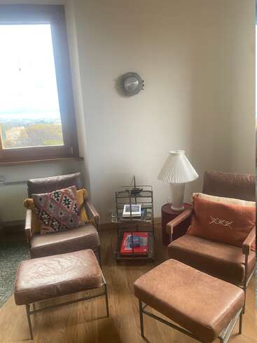 Two brown leather armchairs with matching ottomans sit by a window. Colorful pillows, a side table with books, and a white lamp complete the cozy corner.